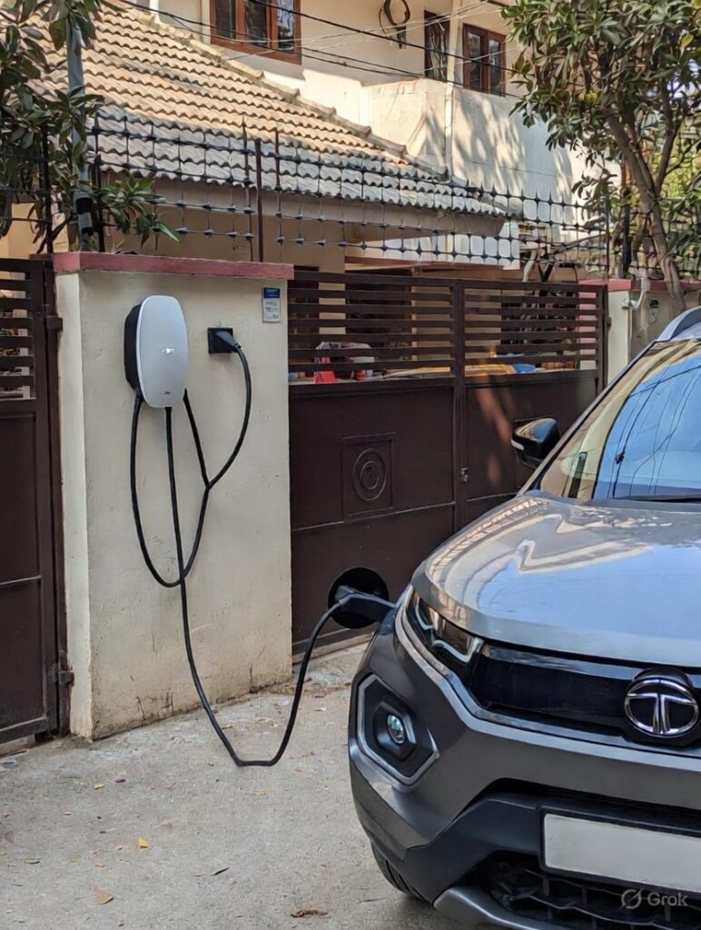 Home EV charger installation in Bengaluru: White 7.4 kW wallbox mounted on a residential compound wall, charging a grey Tata Nexon EV in a typical Indian housing society driveway, showcasing convenient home charging setup 2026
