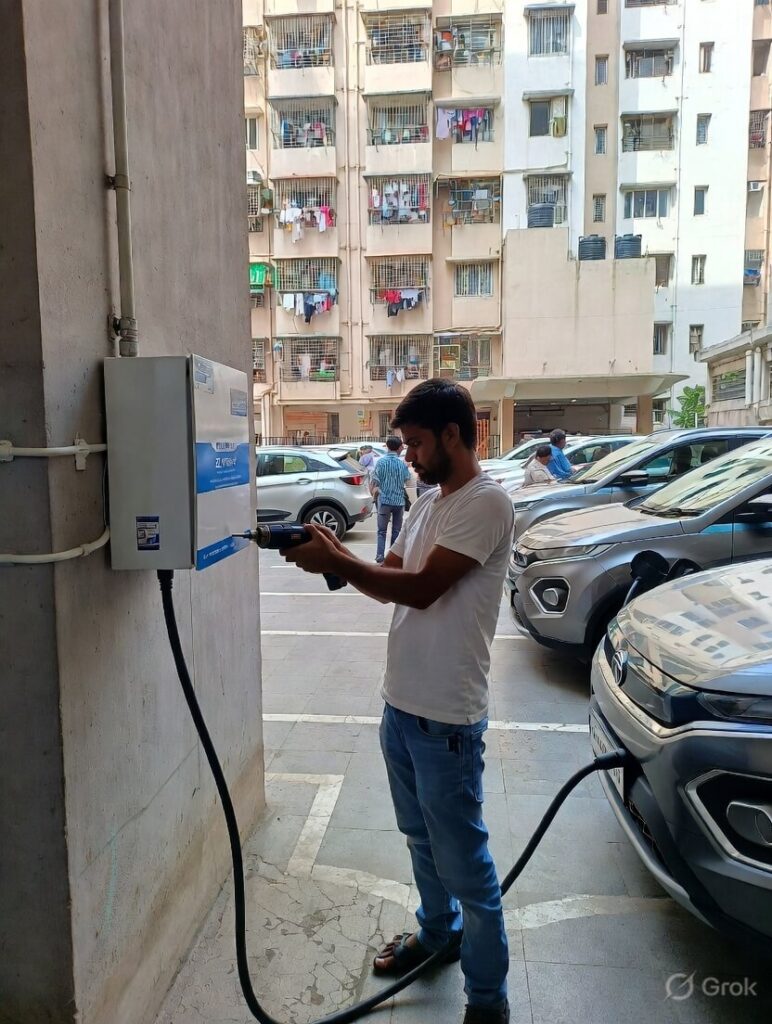 Mumbai resident plugging charging cable into Mahindra XUV400 EV at Zeon 11 kW AC wallbox mounted on apartment building pillar, urban high-rise towers and other cars visible in shared parking area, illustrating reliable home charging for long-distance EV road trips India 2026