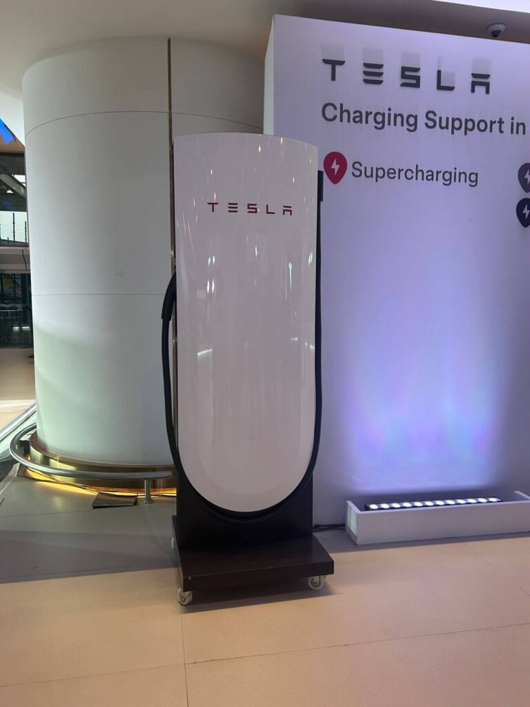 Tesla supercharging support signage at Pune expo April 2026. The charging infrastructure announcement directly impacts any EV charge time calculator 2026 how long to full for your model in US and Europe — with new supercharger corridors planned between Delhi-Jaipur and Ahmedabad-Pune-Bengaluru, 20-80 percent charging drops to 22 minutes for Model Y owners on Indian highways.